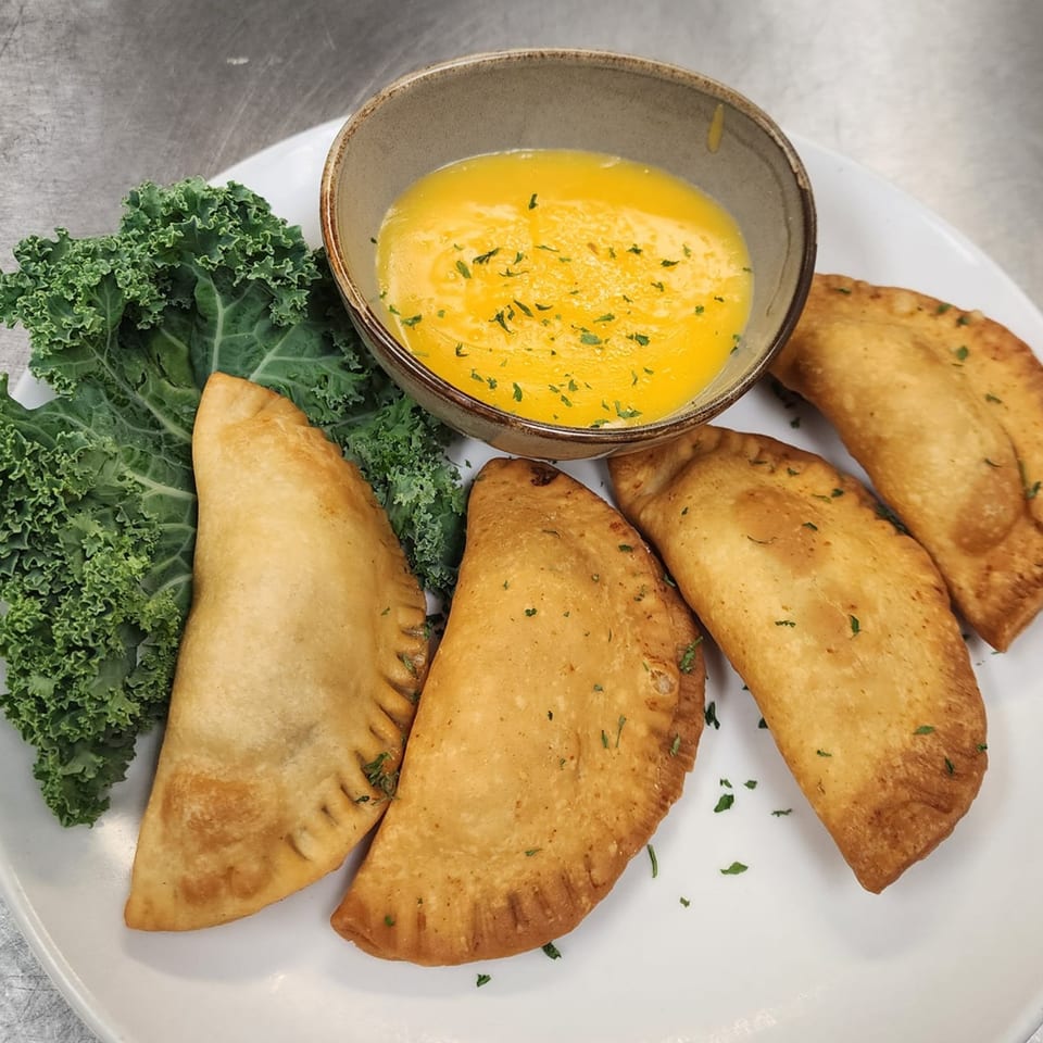Four golden-brown empanadas with a side of bright yellow dipping sauce and green kale.