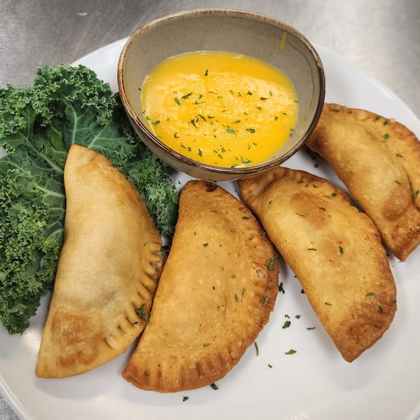Four golden-brown empanadas with kale and a bowl of yellow dipping sauce on a white plate.