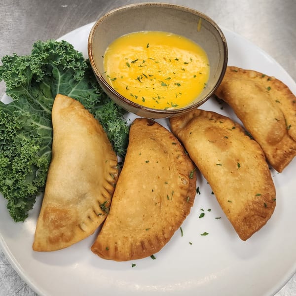 Four golden-brown empanadas with kale and a bowl of melted butter on a white plate.