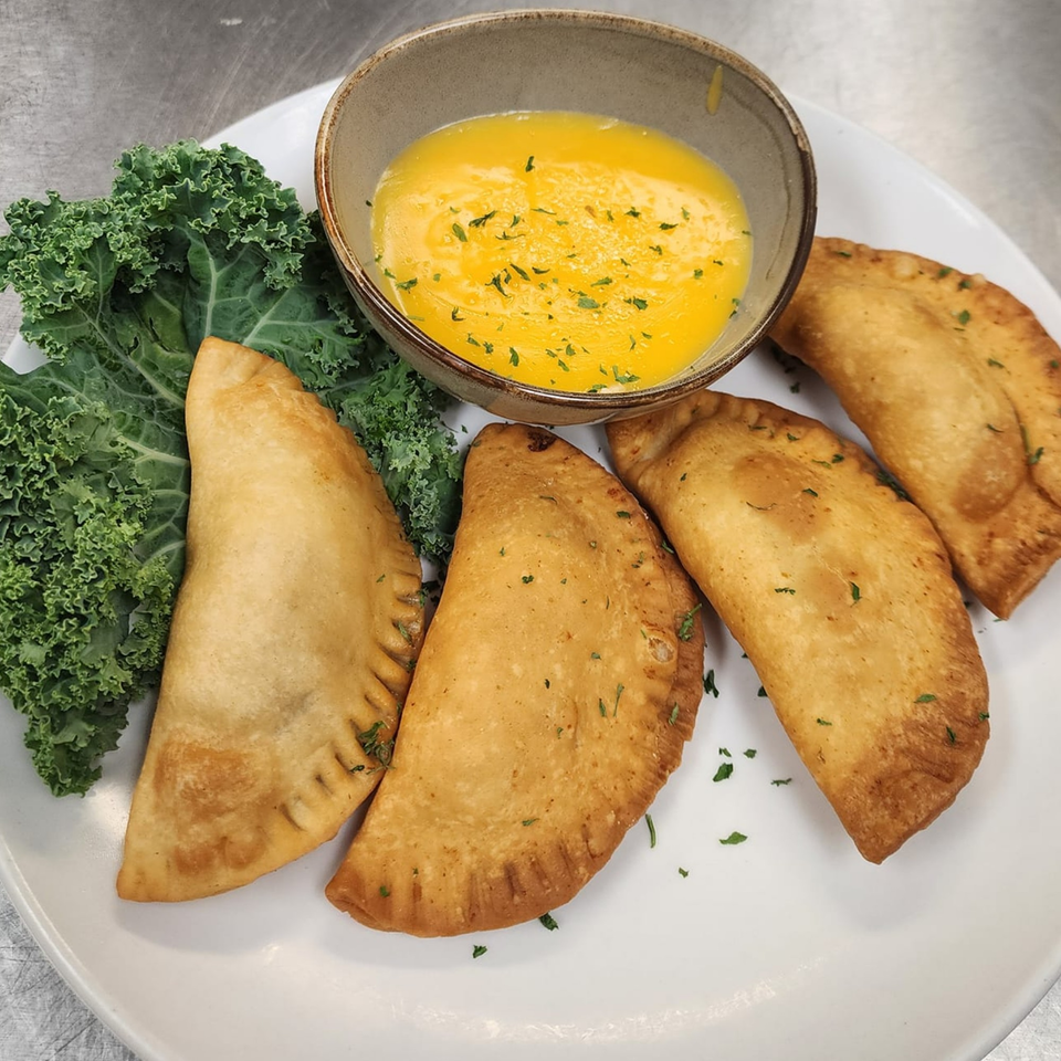 Four golden-brown empanadas with kale and a bowl of melted butter on a white plate.