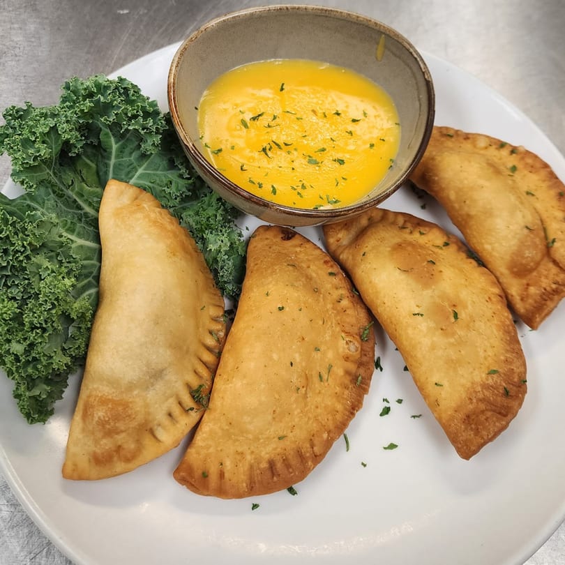 Four golden-brown empanadas with kale and a bowl of melted butter on a white plate.