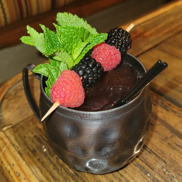 Dark cocktail in a copper mug, garnished with mint, raspberries, and blackberries on a wooden table.