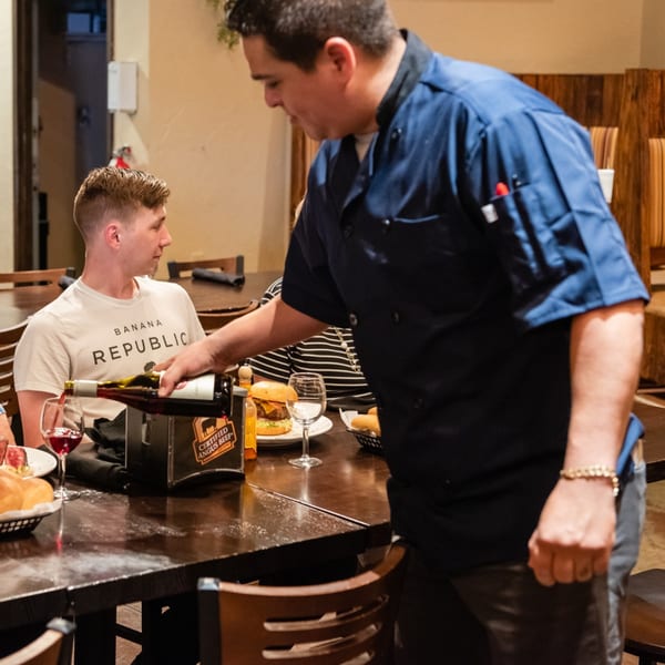 A Christopher's Steakhouse server in a blue chef's coat pours red wine for a customer.